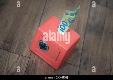 Un primo piano di un conto di cento euro che si stacca da una piccola cassaforte rossa sul pavimento di legno in una casa Foto Stock