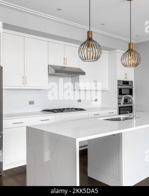 Una grande e lussuosa cucina bianca con una grande isola a cascata, elettrodomestici in acciaio inossidabile, ferramenta in oro e luci dorate appese. Foto Stock