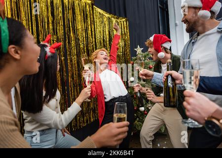 Donna d'affari che balla vicino a colleghi multietnici con champagne e albero di Natale in ufficio Foto Stock