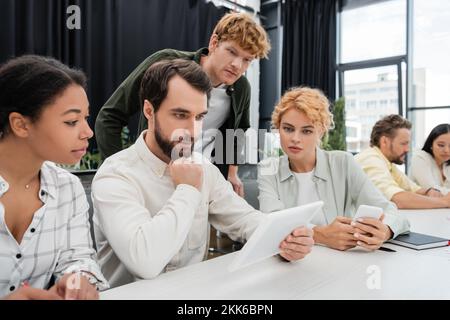 uomo d'affari barbuto che guarda al tablet digitale durante un incontro con colleghi multietnici Foto Stock