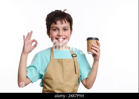 Incredibile barista adolescente in grembiule chef, mostrando segno OK mentre vende bevande calde da asporto in tazza di carta, sorridendo alla macchina fotografica Foto Stock