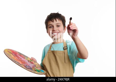 Ragazzo artistico con un pennello e una tavolozza di colori, dipinge su tela immaginaria, sorridendo guardando la macchina fotografica Foto Stock