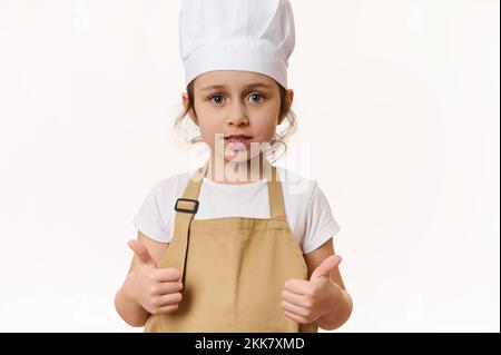 La ragazza carina caucasica che indossa il cappello dello chef e il grembiule beige mostra i pollici in su, guardando la macchina fotografica su sfondo bianco Foto Stock