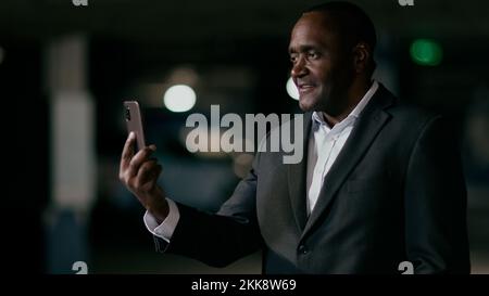 Uomo d'affari afro-americano maturo parla tramite video link utilizzando il telefono cellulare in piedi in stanza oscura puzzled man comunica sulla conferenza online Foto Stock