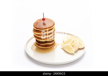 Un mucchio di frittelle ricoperte di miele, servite con una banana affettata su un piatto di ceramica bianca su fondo bianco Foto Stock