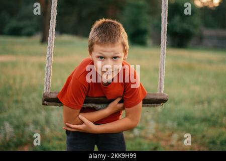 Barefoot Boy 7-10 in t-shirt rossa si appoggia su corda swing in campagna tra pini e guarda in cornice, sorridendo nei raggi del sole tramontare Foto Stock