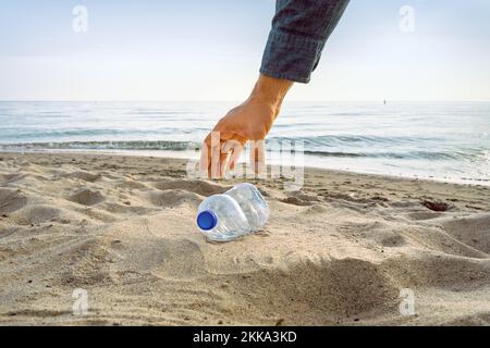 Un braccio a maniche raggiunge per raccogliere una bottiglia vuota su una spiaggia Foto Stock