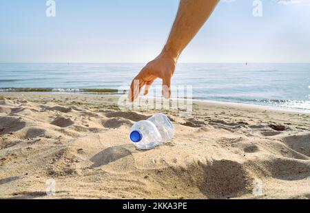 Una mano arriva a prendere una bottiglia d'acqua scartata su una spiaggia Foto Stock