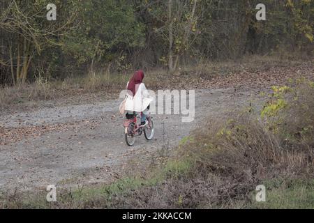 Vecchia donna musulmana in bicicletta lungo una strada con foglie in autunno Foto Stock