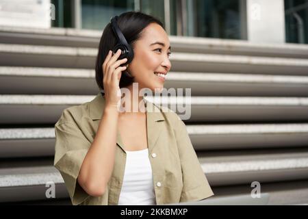 Primo piano ritratto di donna asiatica sorridente ascolta la musica in cuffie, gira con felice espressione del volto, riposa all'aperto Foto Stock