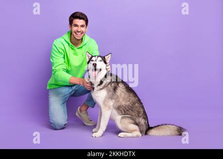 Foto a dimensione piena di allegro divertente soddisfatto ragazzo vestito verde felpa con cappuccio jeans accarezzare il suo cane Husky isolato su sfondo di colore viola Foto Stock