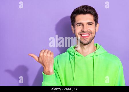 Foto di bel ragazzo positivo con brunet hairdo vestito felpa verde con cappuccio che indica spazio vuoto isolato su sfondo di colore viola Foto Stock