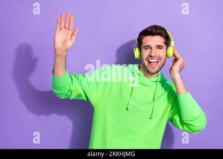 Ritratto di ottimista bello attraente ragazzo brunet hairdo verde con cappuccio cuffie touch alto cinque isolati su sfondo di colore viola Foto Stock