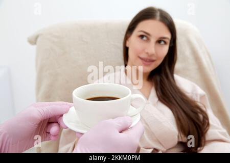 Beautician che serve una tazza di caffè al suo cliente dopo il trattamento del corpo nel salone di bellezza. Rilassatevi con le bevande su un letto cosmetico. Bellezza, piacere Foto Stock