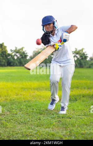 Giocatore di cricket femminile che indossa un equipaggiamento protettivo e colpisce il pallone con un pipistrello sul campo Foto Stock