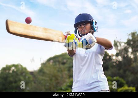 Giocatore di cricket femminile che indossa un equipaggiamento protettivo e colpisce il pallone con un pipistrello sul campo Foto Stock