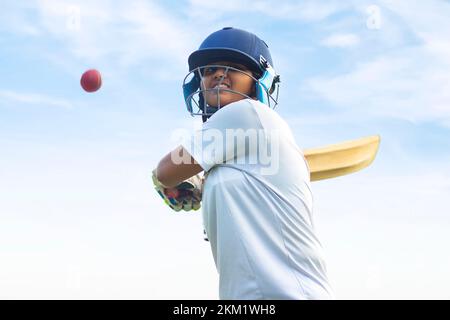 Giocatore di cricket femminile che indossa un equipaggiamento protettivo e colpisce il pallone con un pipistrello sul campo Foto Stock