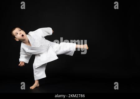 Un ragazzino in un kimono pratica il karate su sfondo nero, calciando in avanti. Foto Stock