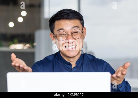 Foto ravvicinata di un uomo d'affari asiatico di successo che parla in remoto durante una videochiamata, un capo maschio in occhiali che guarda lo schermo del computer portatile sorridendo e parlando amichevole, un programmatore che lavora all'interno dell'ufficio. Foto Stock