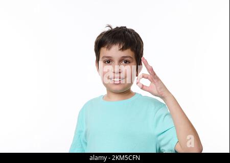 Ragazzo caucasico dai capelli scuri che indossa una t-shirt casual, sorridendo e mostrando un segno ok guardando la macchina fotografica isolata su una parete bianca Foto Stock