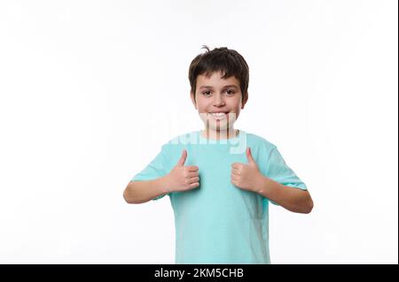 Ritratto di un simpatico ragazzo adolescente in t-shirt blu casual, che mostra il pollice in alto nello spazio di copia per il testo, su sfondo bianco Foto Stock