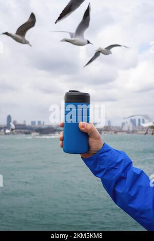 Mano maschile che tiene una tazza di caffè da viaggio thermos sullo sfondo di acqua limpida di un mare con una tonalità turchese. Foto Stock