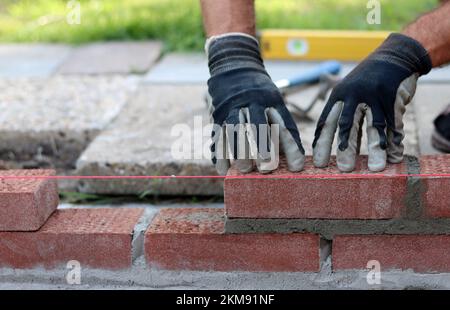 Costruire un muro di mattoni rossi. Mani maschili che tengono cazzuola in mattoni e strumento di misurazione della livella. Lavori di muratura nei dettagli. Foto ravvicinata del cantiere. Foto Stock