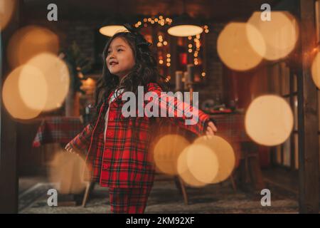 Attiva ragazza bambino giappone con elegante modello di capelli brunette lunga posa a casa in bokeh ghirlands noel. Asian piccolo fashionista in rosso plaid pajama danza Foto Stock