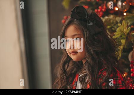 Felice giappone piccola ragazza sognatore guardando via miracolo di attesa da Santa posa vicino a noel albero. Capelli ricci neri asiatici capretto in abito rosso celebrato a plaid Foto Stock