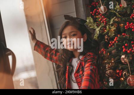 Felice giappone piccola ragazza sognatore guardando attraverso la finestra e miracolo in attesa da Santa posa vicino a noel albero. Bambino asiatico in abito rosso plaid festeggiamento Foto Stock