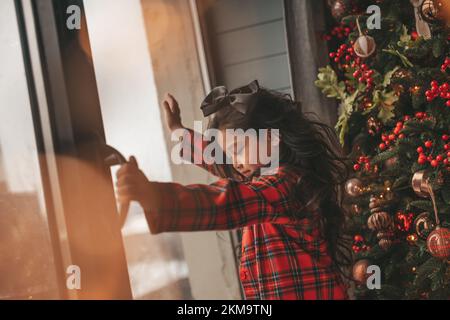 Felice giappone piccola ragazza sognatore guardando attraverso la finestra e miracolo in attesa da Santa posa vicino a noel albero. Bambino asiatico in abito rosso plaid festeggiamento Foto Stock