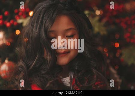 Felice giappone piccola ragazza sognatore guardando via miracolo di attesa da Santa posa vicino a noel albero. Capelli ricci neri asiatici capretto in abito rosso celebrato a plaid Foto Stock