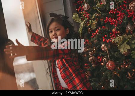 Felice giappone piccola ragazza sognatore guardando attraverso la finestra e miracolo in attesa da Santa posa vicino a noel albero. Bambino asiatico in abito rosso plaid festeggiamento Foto Stock