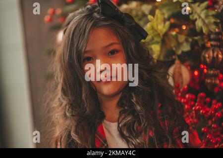 Felice giappone piccola ragazza sognatore guardando via miracolo di attesa da Santa posa vicino a noel albero. Capelli ricci neri asiatici capretto in abito rosso celebrato a plaid Foto Stock