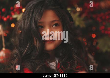 Felice giappone piccola ragazza sognatore guardando via miracolo di attesa da Santa posa vicino a noel albero. Capelli ricci neri asiatici capretto in abito rosso celebrato a plaid Foto Stock