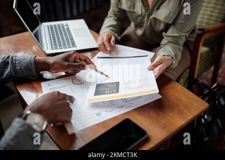 Mano di un giovane uomo nero con penna che indica il documento finanziario mentre spiega i dati a una collega durante la discussione Foto Stock