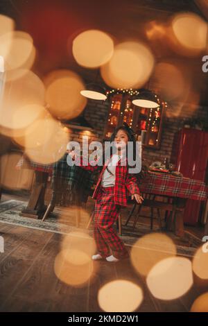 Attiva ragazza bambino giappone con elegante modello di capelli brunette lunga posa a casa in bokeh ghirlands noel. Asian piccolo fashionista in rosso plaid pajama danza Foto Stock