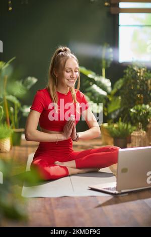 giovane donna in abiti rossi per il fitness con un computer portatile che fa yoga e videochattare nella moderna casa verde. Foto Stock