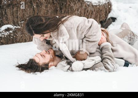 Giovane coppia ragazza ragazzo a piedi vicino fienile con pattini. Inverno freddo innevato in campagna. Pattinare, baciare, divertirsi, ridere in abiti eleganti, pelliccia Foto Stock