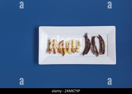 Un piatto rettangolare in ceramica bianca con patate al rosmarino e acciughe al forno in fila pronto per servire su uno sfondo blu studio con spazio copia. Foto Stock