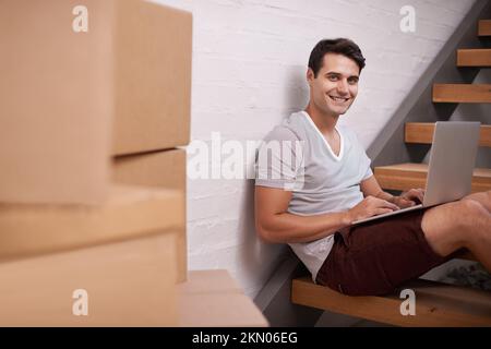 Invio dell'ultima e-mail nella mia vecchia casa. Un giovane uomo felice che usa il suo computer a casa tra le scatole imballate. Foto Stock