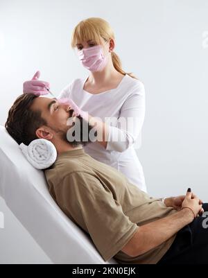 Primo piano dell'uomo bearded che ottiene l'iniezione di bellezza alla clinica estetica. Medico che inietta il riempitivo anti-invecchiamento nel bel viso maschile Foto Stock