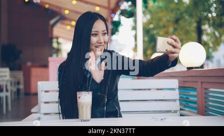 Happy Asian lady sta parlando con gli amici online facendo videochiamata utilizzando lo smartphone seduto in un bar all'aperto da solo. La ragazza sta parlando, toccando lunghi capelli neri e sorridendo. Foto Stock