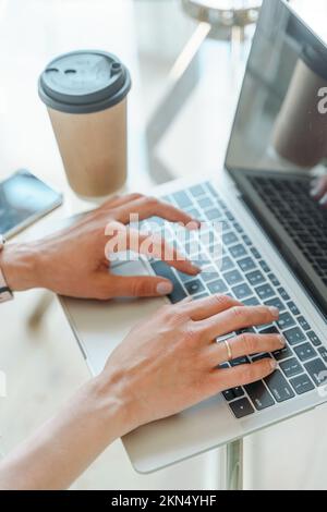 Mani che scrivono sulla tastiera di un computer sopra un tavolo da ufficio bianco con una tazza di caffè e forniture, vista dall'alto. Foto Stock