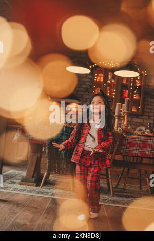 Attiva ragazza bambino giappone con elegante modello di capelli brunette lunga posa a casa in bokeh ghirlands noel. Asian piccolo fashionista in rosso plaid pajama danza Foto Stock