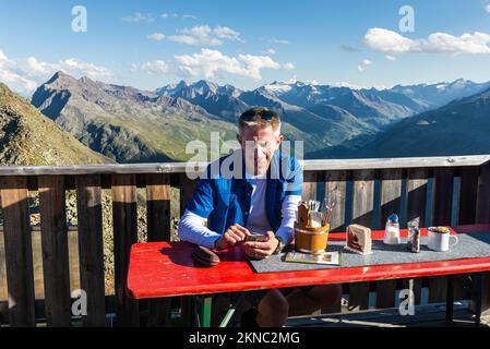 Un escursionista si siede sulla terrazza della capanna montana Brunnenkogelhaus e guarda al suo cellulare di fronte alle Alpi di Ötztal e alla valle di Gurgl, Austria Foto Stock