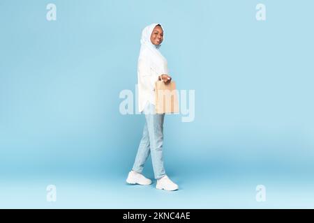 Concetto di vendita stagionale. Foto a tutta lunghezza di una donna musulmana nera in hijab camminando con la borsa della spesa su sfondo blu Foto Stock