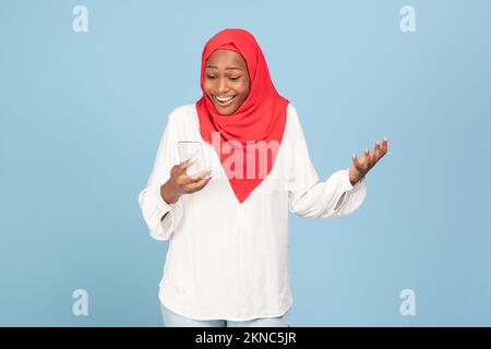 Donna musulmana afro-americana overjoyed che guarda smartphone in eccitazione, posa su sfondo blu studio Foto Stock