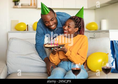 Compleanno del marito. Moglie afro-americana sorprende il marito con la torta di compleanno, l'uomo che si gode b-giorno celebrazione a casa Foto Stock