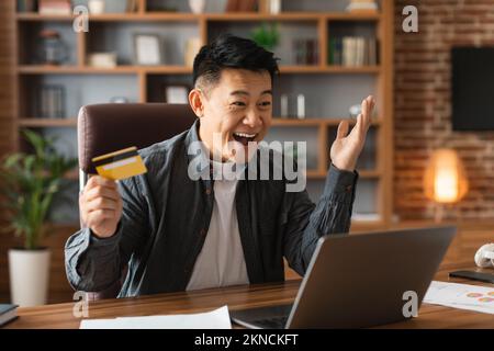 Allegro sorpreso eccitato maschio cinese maturo con bocca aperta tenere carta di credito e guardare il computer Foto Stock